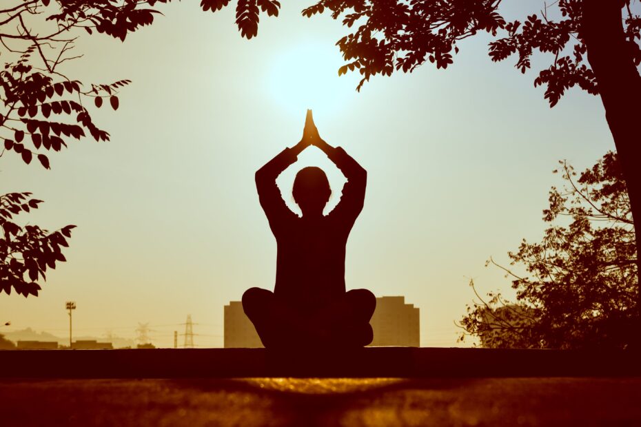 A silhouette of someone sitting in the lotus position during sunrise with an industrial skyline behind them